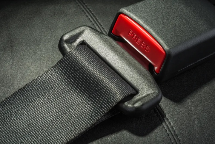 Close view of woven seat belt webbing threaded through a latch plate beside a red release button on the buckle